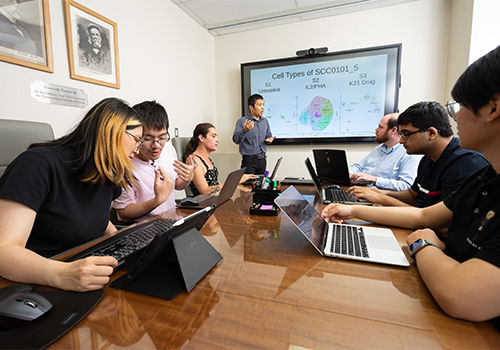 seven students in a dry lab room on laptops sitting around a table watching a presenter standing by a screen