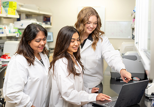 three women in white lab coats smiling and staring at laptop screen in a lab