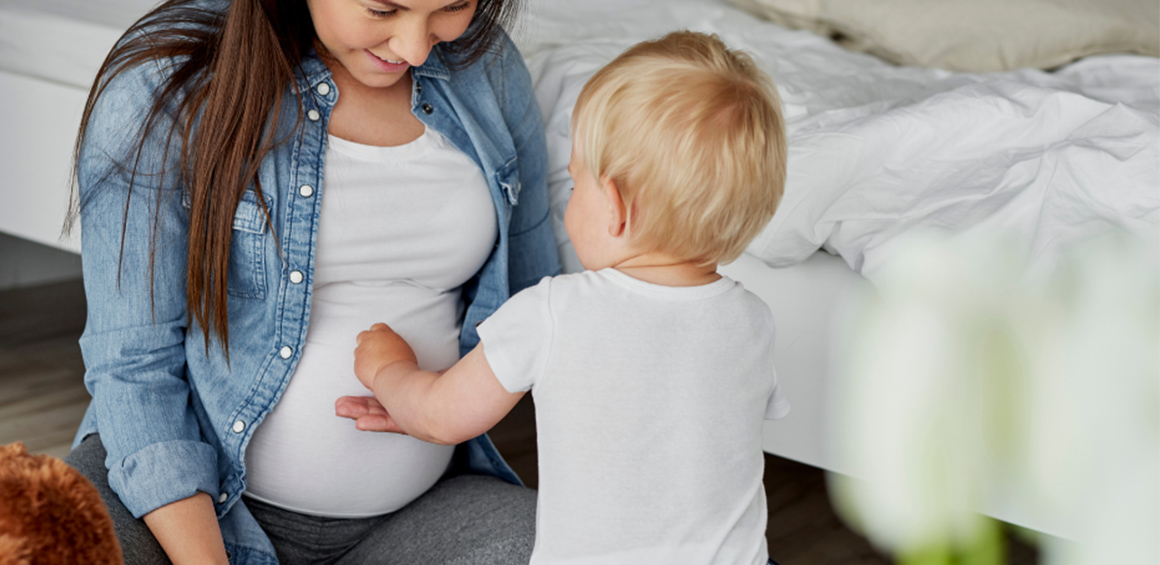 Pregnant mother sitting smiling with an infant in front of her placing his hand over her stomach